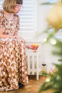 Low plain Mercury 6 Column 360mm cast iron radiator in off-white on feet, brass valve, under shuttered window on parquet, woman in gold sequins beside it.
