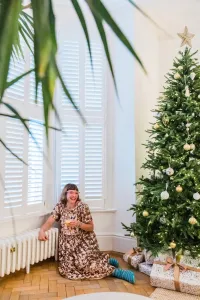 A white Mercury 6 column cast iron radiator on feet beneath shuttered windows, with a woman seated on a parquet floor beside a Christmas tree.