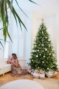 A low white Mercury cast iron radiator on feet beneath shuttered bay windows, beside a woman seated on parquet near a decorated Christmas tree.