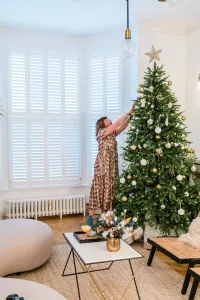 A low white Mercury cast iron radiator under a shuttered bay window, beside a woman decorating a Christmas tree on a herringbone floor.