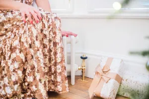 Low white Mercury 6 Column 360mm cast iron radiator under a window with a brass valve, wrapped gifts on a parquet floor and a woman in a sequin dress.