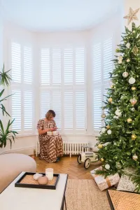 A low white Mercury 6 Column cast iron radiator beneath a shuttered bay window, with a woman wrapping gifts beside a Christmas tree.