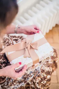 Hands holding a gift tied with rose-gold ribbon over a sequinned skirt, a low white Mercury cast iron radiator blurred behind on a parquet floor.