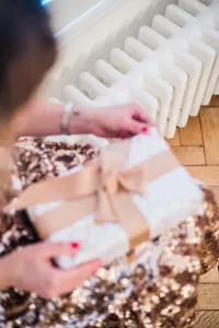 A woman in a gold sequin top unties the ribbon on a white gift beside a low white Mercury 6 Column cast iron radiator on a parquet floor.