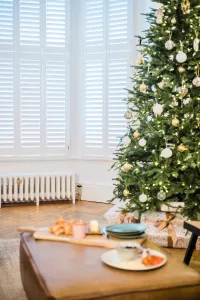 A low white Mercury 6 column cast iron radiator with a brass valve beneath white shuttered bay windows, beside a decorated Christmas tree.