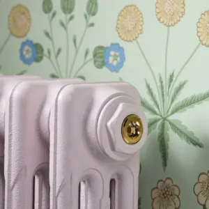 Close-up of a plain Mercury cast iron radiator in pale lilac with a Polished Brass BVA1 bleed valve, against green floral wallpaper.