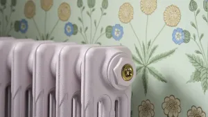 Close-up of a plain Mercury cast iron radiator painted pale lilac, with a Polished Brass BVA1 bleed valve, against floral green wallpaper.