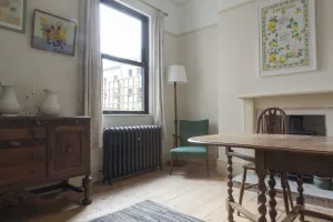 Ornate Rococo II 760mm cast iron radiator in Astral Grey Dark on feet below a window, in a dining room with wooden floorboards and a green armchair.