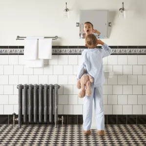 Ornate Rococo II cast iron radiator in Etch-Coated Polish on feet with valves, in a white-tiled bathroom as a boy brushes his teeth.