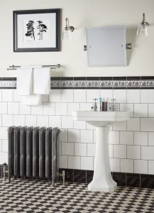Ornate Rococo II cast iron radiator in Etch-Coated Polish on feet with angled valves, beside a white pedestal basin on a chequered floor.