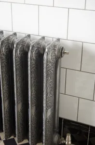 Ornate Rococo II cast iron radiator in Etch-Coated Polish with a chrome bleed valve and angled valve, against white metro tiles above a chequered floor.