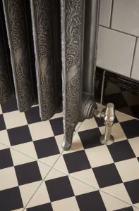 Close-up of an ornate Rococo II cast iron radiator in Etch-Coated Polish on feet, with a satin nickel valve, on black and cream chequered tiles.