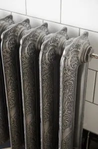 Close-up of ornate Rococo II cast iron radiator sections in Etch-Coated Polish, with a valve fitting, against white wall tiles.