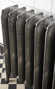 Close view of an ornate Rococo II 760mm cast iron radiator in Etch-Coated Polish on feet, against white tiles above a black and white chequered floor.