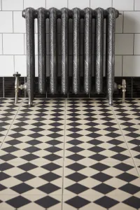 Ornate Rococo II 760mm cast iron radiator in Etch-Coated Polish on feet, with angled valves, against white tiles above a black and cream chequered floor.