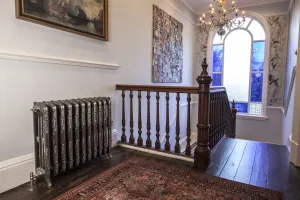 Ornate Rococo cast iron radiator in Full Polish on feet with a valve, on a landing with wooden banister, rug and arched window.
