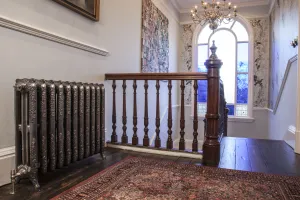 Ornate Rococo III cast iron radiator in Full Polish on feet, with a valve, on a landing with a wooden banister, rug and arched window.
