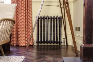 Ornate Rococo III cast iron radiator in Full Polish on feet, with angled valves, on a herringbone floor beside a patterned orange curtain.
