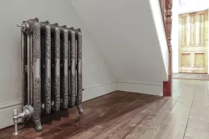 Ornate Rococo III cast iron radiator in Full Polish on feet, with polished angled valves, under a staircase on wooden floorboards.
