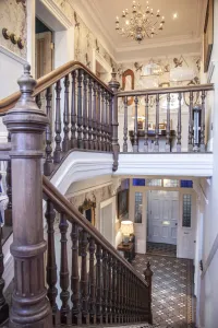 Victorian hallway with a carved wooden staircase, patterned wallpaper, crystal chandelier and geometric tiled floor leading to a panelled front door.