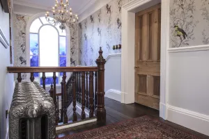 Ornate Rococo III cast iron radiator in Full Polish beside a carved wooden banister on a landing with bird wallpaper and an arched window.