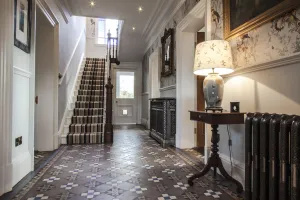Ornate Rococo III cast iron radiator in Full Polish beside a lamp table in a hallway with Victorian patterned tiles and a striped stair runner.