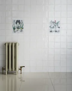 Ornate Rococo III cast iron radiator in Minster White on feet, with brass valves, against white tiles with botanical panels and a glossy floor.