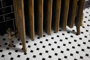 Ornate Rococo cast iron radiator in Natural Brass on feet, with an angled brass valve, on white octagonal floor tiles with black insets.