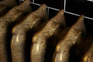 Close-up of ornate Rococo III cast iron radiator columns with scrolled relief in Natural Brass, against black tiles with white grout.