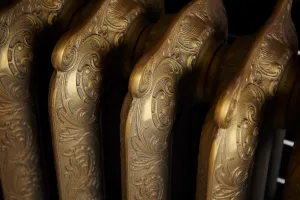 Close-up of the ornate scrolled columns of a Rococo III cast iron radiator in Natural Brass, set against a dark background.