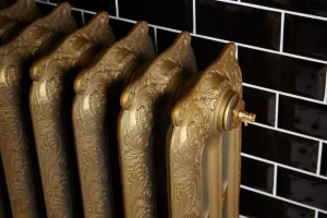 Close-up of an ornate Rococo cast iron radiator in Natural Brass with scrolled columns and a brass bleed valve, against black metro tiles.