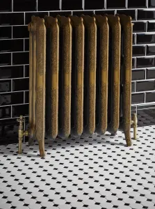 Ornate Rococo III cast iron radiator in Natural Brass on feet, with brass angled valves, against black metro tiles on a black and white mosaic floor.