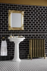 Ornate Rococo III cast iron radiator in Natural Brass on feet with brass valves, beside a pedestal basin in a black metro-tiled bathroom.