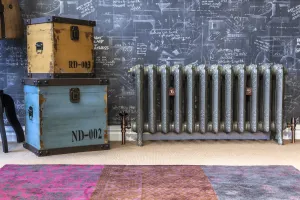 Ornate Rococo II cast iron radiator on feet in a grey finish with copper valves, beside stacked trunks against chalkboard-print wallpaper.