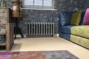 Ornate Rococo II 510mm cast iron radiator on feet in a metallic grey finish with copper valves, under a window beside a blue leather sofa.