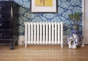 Ornate Rococo II 510mm cast iron radiator in Farrow & Ball Wimborne White on feet, with nickel valves, against blue tree-print wallpaper on floorboards.