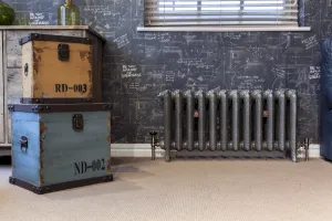 Ornate Rococo II cast iron radiator on feet in a metallic pewter finish with copper valves, below a window beside vintage trunks on carpet.