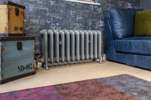 Ornate Rococo II cast iron radiator in a grey finish on feet with copper valves, beneath a chalkboard wall beside a blue leather sofa.
