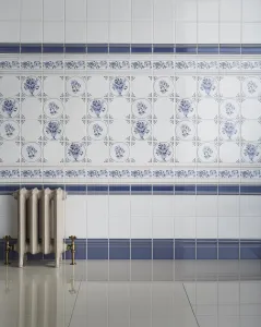 A small ornate Rococo II cast iron radiator in cream on feet, with brass valves, against white and blue Delft-style floral wall tiles.
