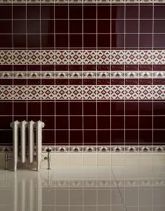A short ornate Rococo II cast iron radiator in a cream finish on feet, with chrome valves, against glossy burgundy and patterned wall tiles.