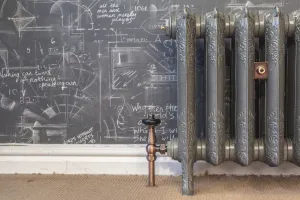 Ornate Rococo II cast iron radiator in a dark grey metallic finish on feet, with a copper angled valve, before a chalk-covered wall.