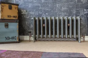Ornate Rococo II 510mm cast iron radiator in metallic grey on feet, with copper valves and wall stays, against chalkboard wallpaper.