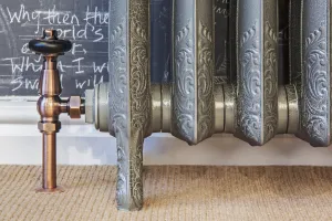 Close-up of an ornate Rococo II cast iron radiator on feet in a silver crackle finish, with a copper angled valve, carpet and chalkboard wall.