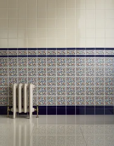 Ornate Rococo II 510mm cast iron radiator in an off-white finish with brass valves, against blue floral wall tiles on a glossy floor.