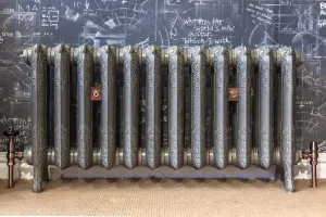 Ornate Rococo II cast iron radiator on feet in a metallic silver finish, with copper valves and wall stays, against a chalkboard wall on sisal flooring.