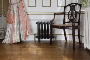 A low ornate Rococo II cast iron radiator in Matt Black on feet with brass valves, beside a carved armchair and curtains on a wooden floor.
