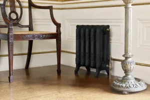 Ornate Rococo II cast iron radiator in Matt Black on feet, with a brass valve, between a carved chair and a painted lamp stand on oak boards.