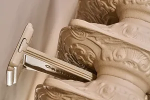 Close-up of a Castrads-branded wall stay fixed to a wall, holding the ornate scrolled sections of a Rococo II radiator in a pale stone finish.