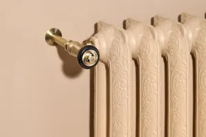 Close-up of an ornate Rococo II cast iron radiator in Farrow & Ball Planter No. G5 with an antique brass valve against a beige wall.