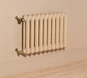 Ornate wall-hung Rococo II cast iron radiator in a cream finish with brass angled valves, on a beige wall above a skirting board and oak floor.